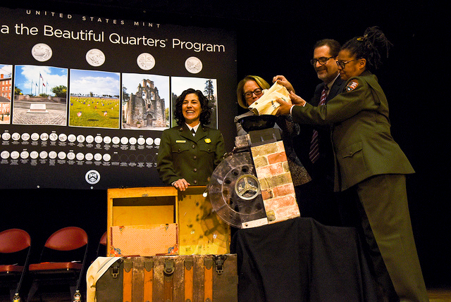 three women and a man pour coins from a bag into a wheel that drops them into a wood box