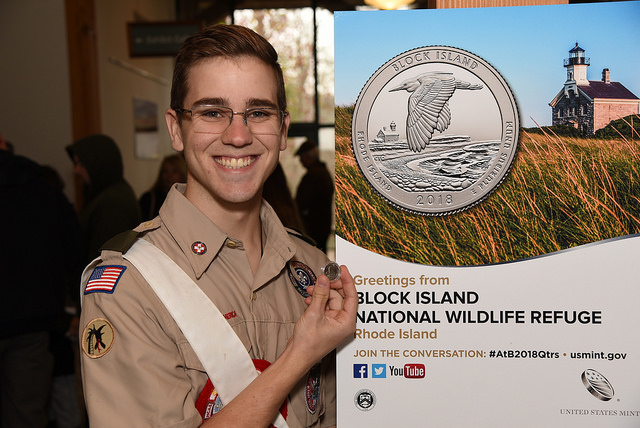 an Eagle Scout holds a quarter and stands next to a Block Island National Wildlife Refuge event poster with an image of the quarter and lighthouse