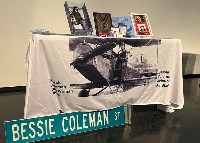 Bessie Coleman doll, books, and photos on a table with a tablecloth printed with an image of Bessie Coleman and her plane